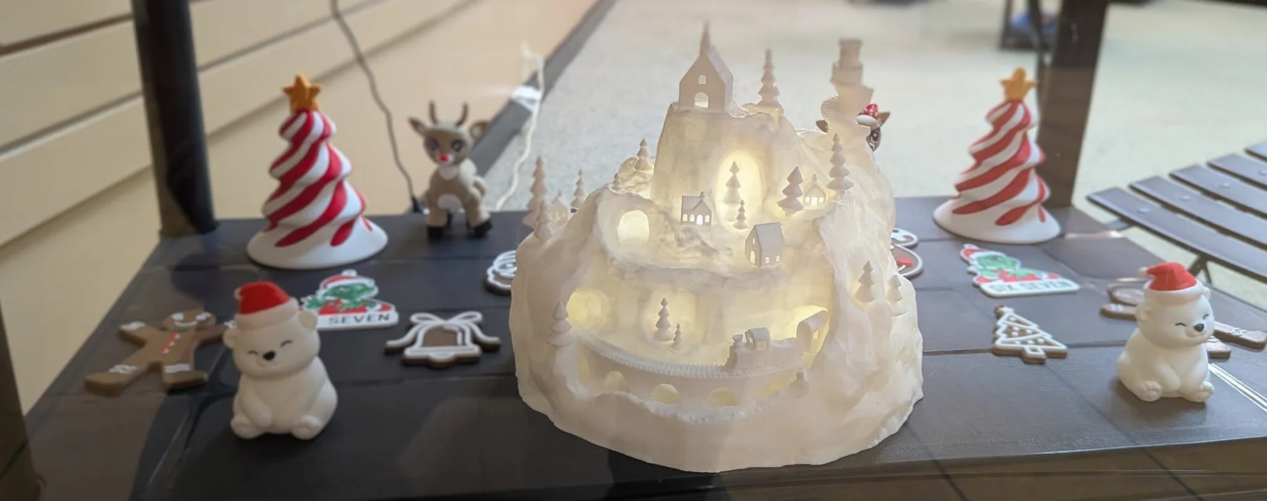 A glowing 3D-printed light-up village display with a snowy hillside and tiny lit windows.