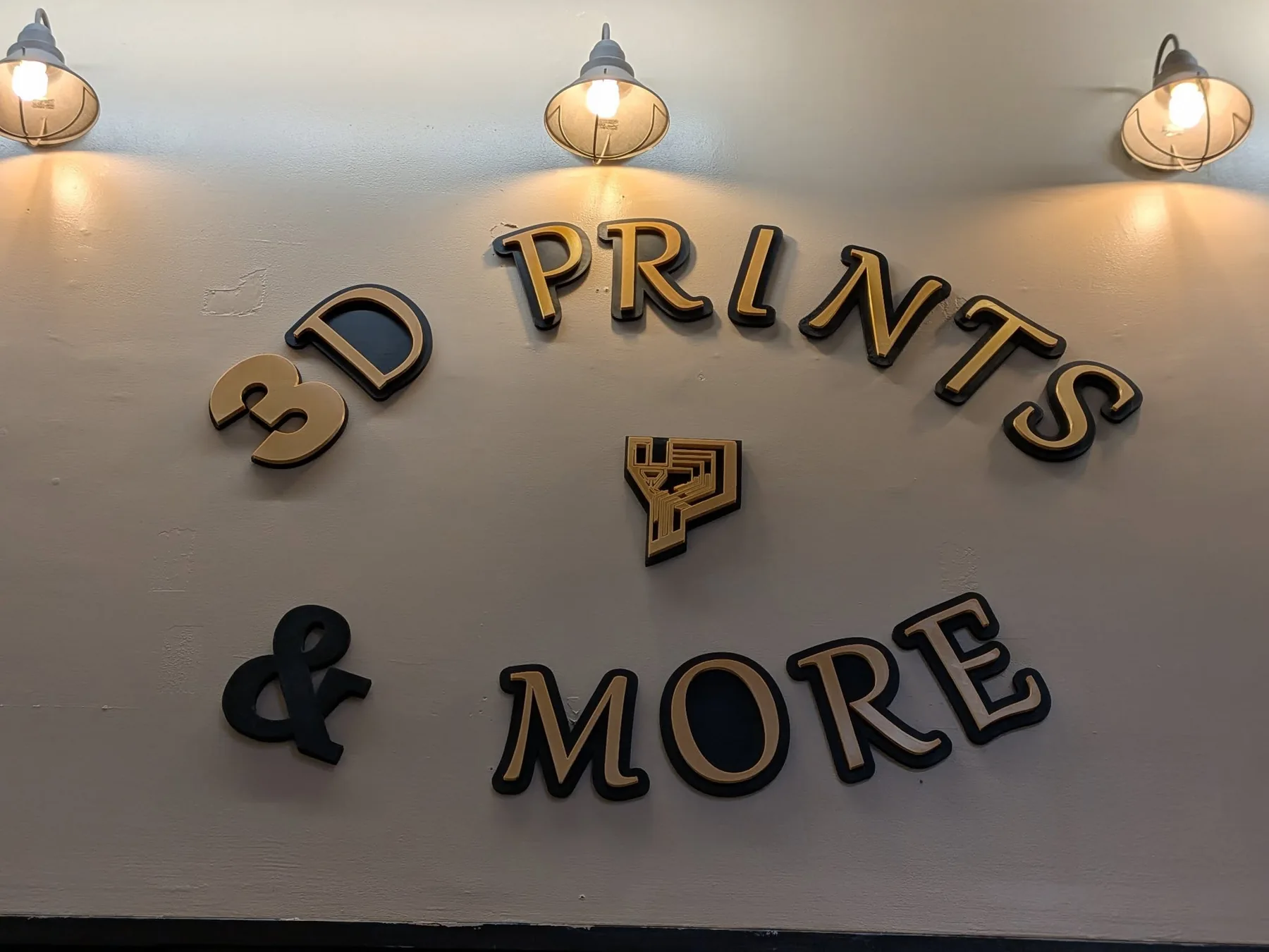 Close-up of the gold 3D Prints and More wall sign with the shop's logo mark.
